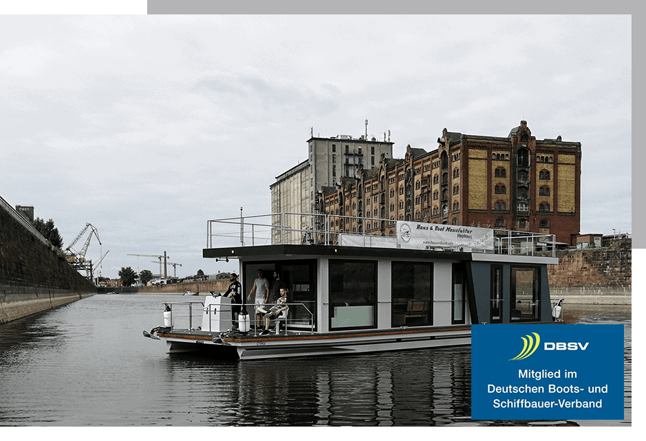 Ein modernes Hausboot fährt auf einem Fluss vor einem alten Backsteingebäude.